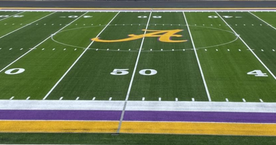 Turf football field with a purple and gold sideline and a gold A in the center