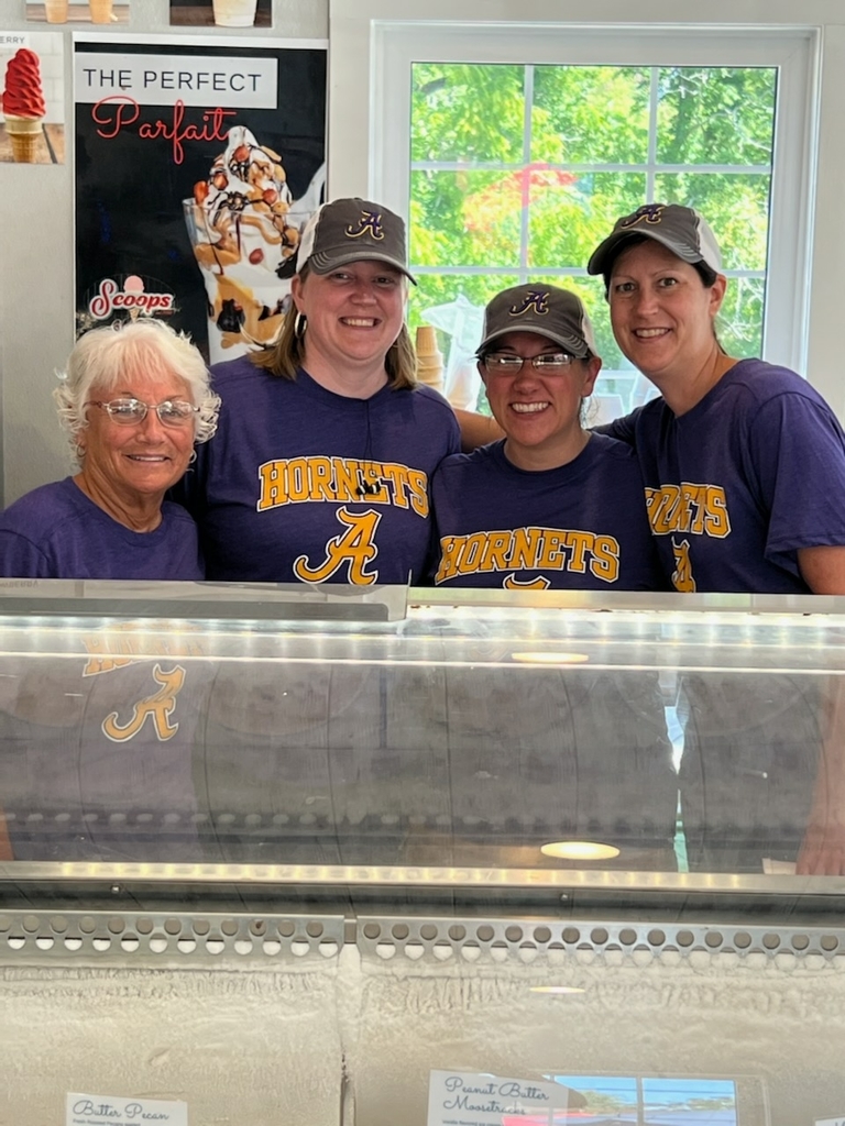 Four women in purple and gold Angola Hornets T shirts pose with smiles behind an ice cream freezer