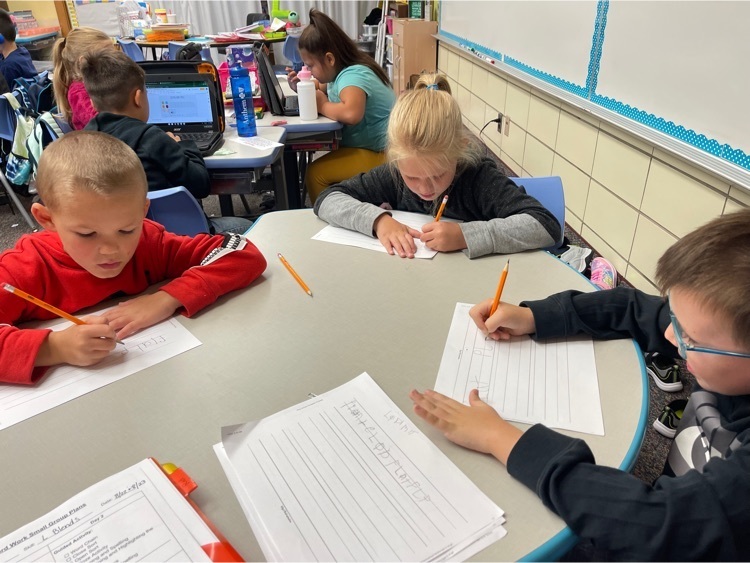 Students sitting in a small group writing words on paper with a pencil. 
